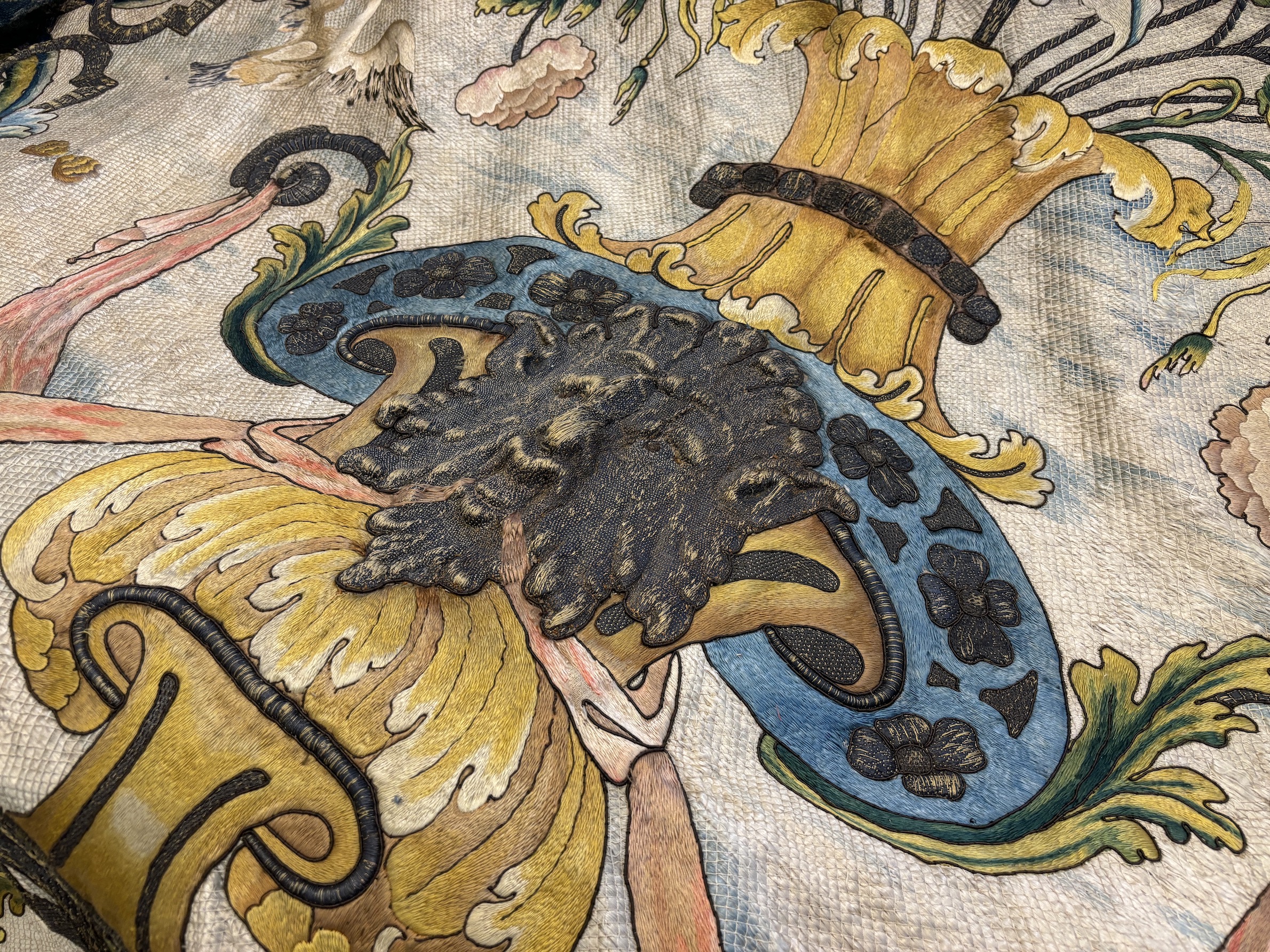 A large early 18th century possibly French polychrome and gold metallic silk embroidered wall hanging, with scrolling vine and floral border. The central motifs embroidered with an elaborate metallic thread cartouche fra